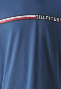Modrá látka so horizontálnym pleteným prúžkom v červenej, bielej a čiernej farbe s čiastočne viditeľným textom "HILFIGER".