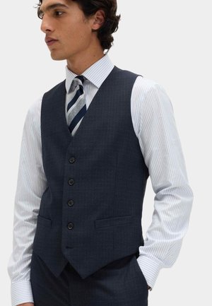 Man draagt een donkerblauw knoopvest over een wit overhemd met dunne blauwe strepen, gecombineerd met een gestreepte marineblauwe en grijze stropdas.