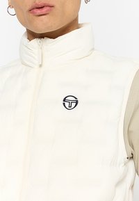 Chaleco acolchado blanco sin mangas con cuello alto, cierre de cremallera y textura acolchada. Presenta un pequeño logo negro en la zona del pecho.