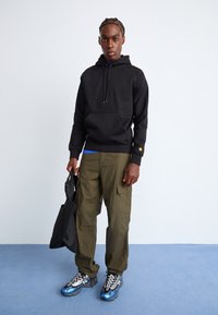 Zwarte hoodie met kangoeroezak, olijfgroene cargobroek en blauwe sneakers met zilveren accenten. Houdt een zwarte tote bag vast.