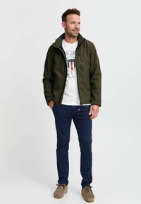 Olivgrüne Softshell-Jacke mit Reißverschluss, kombiniert mit einem weißen Grafik-T-Shirt, navyblauen Hosen und hellbraunen Wildlederschuhen.