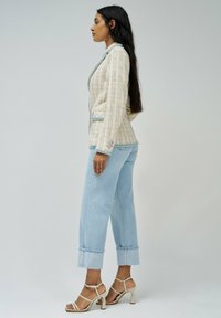 Blazer de tweed a medida en crema con acentos pastel, con cuello de pico y bordes deshilachados. Combinado con jeans cortos de lavado claro y sandalias de tacón beiges.