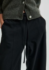 Šedý pletený kardigán s rebrovanou textúrou a gombíkovými detailmi, skombinovaný s čiernymi nohavicami s elastickým pásom a hladkým materiálom.