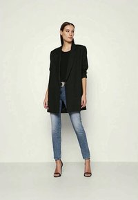 Žena v čiernej oversize saku, čiernom tope, svetlom modrých skinny džínsach a čiernych vysokých sandáloch stojí proti jednoduchému pozadiu.