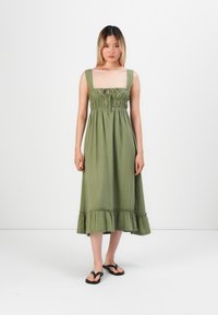 Maxikjole i salviegrønn stoff, med firkantet halsutskjæring, smocket overdel og volangkant. Brukt med svarte flip-flops.