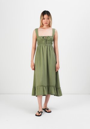 Maxi dress in sage green fabric, featuring a square neckline, smocked bodice, and ruffled hem. Worn with black flip-flops.