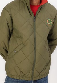 Olivengrøn quiltet jakke med zip-front, rød indre krave, GANT-logo i grøn og orange samt sidelommer med knapdetaljer.