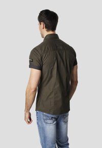 Camisa de manga corta color verde oliva con cuello con solapa, mangas remangadas y un parche con logo en el brazo izquierdo, en un corte casual.