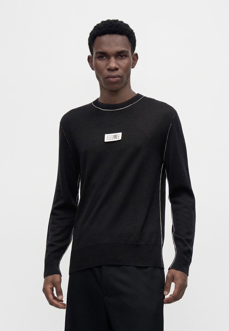 Mann trägt schwarzen Langarmpullover mit weißen Steppnähten und kleinem rechteckigem Logo-Patch auf der Brust vor einfarbigem Hintergrund.