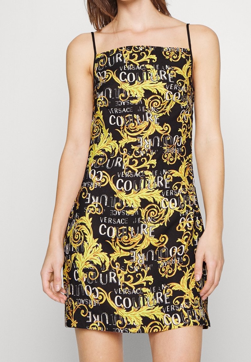 Femme portant une mini-robe noire à fines bretelles ornée de motifs baroques jaunes et texte blanc "Versace Jeans Couture".