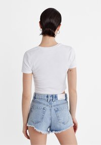 Mujer con cabello corto y mojado, que lleva pendientes de aro plateados, una blusa blanca ajustada de manga corta y unos pantalones cortos de mezclilla de tiro alto deshilachados de color azul claro.