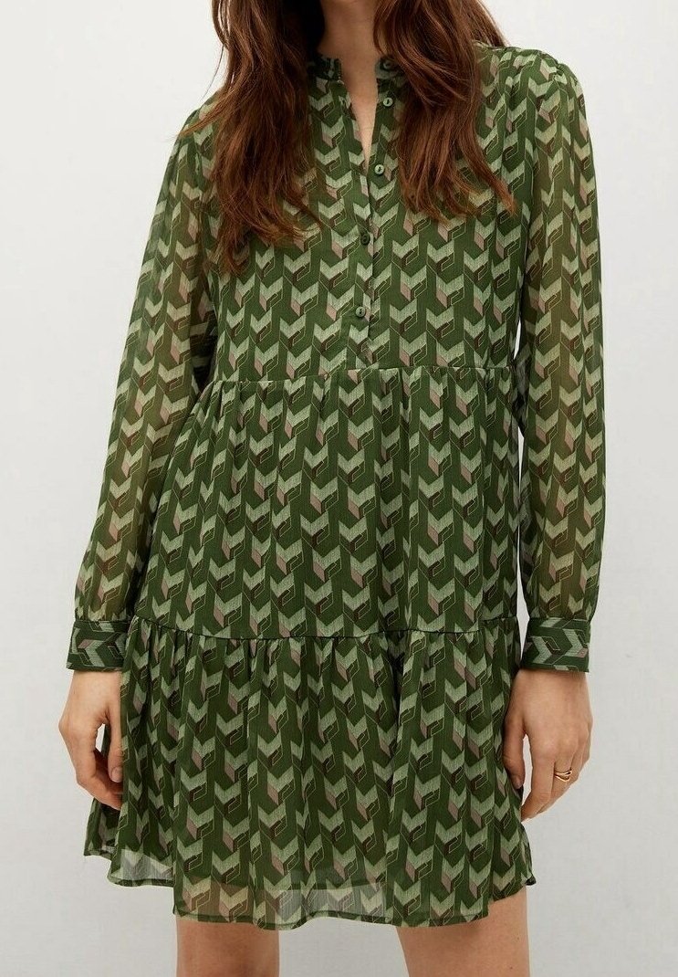 Femme portant une robe verte à motif géométrique, manches longues, boutonnée devant, jupe à volants et coupe ample.