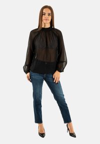 Blusa negra semitransparente con estampado de lunares, cuello alto y mangas largas abullonadas. Combinada con vaqueros azules y zapatos de tacón negro.