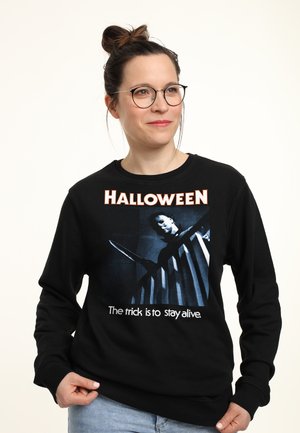 Donna con occhiali che indossa una felpa nera di Halloween che mostra una figura mascherata che tiene un coltello sulle scale.