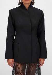 Musta räätälöity bleiseri, jossa on korkea kaulus, rakenteelliset harteet ja kaksi etutaskua. Sujuva kangas, joka on muotoiltu istuvaksi.