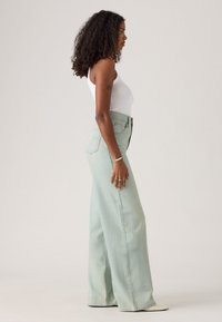 Light green wide-leg jeans made of denim, featuring a high waist and a subtle faded texture, paired with a white fitted tank top.
