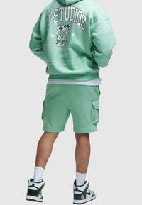 Homme portant un sweat à capuche vert menthe et un short cargo avec un texte graphique au dos, des chaussettes blanches et des baskets noires et blanches, debout devant un fond uni.