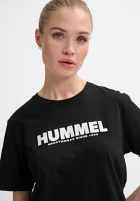 Femme aux cheveux blonds attachés, portant un t-shirt noir "HUMMEL Sportswear depuis 1923", debout devant un fond blanc uni.