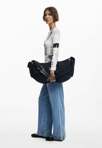 Marinblå axelväska med en texturerad yta, stor storlek och justerbar rem, hålls av en person som bär en grafisk långärmad tröja och vida jeans.