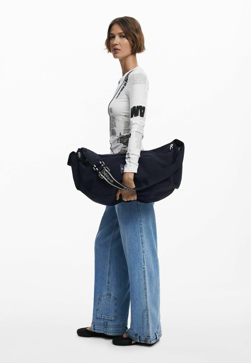 Marinblå axelväska med en texturerad yta, stor storlek och justerbar rem, hålls av en person som bär en grafisk långärmad tröja och vida jeans.