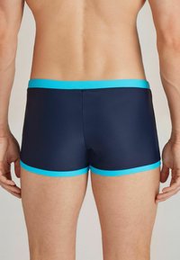 Marinefarbene Schwimmshorts mit einem figurbetonten Design, ausgestattet mit einem leuchtend blauen Gummibund und Saum. Glatte Textur, hüftlanger Schnitt.
