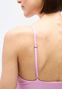 Górna część pleców kobiety z krótkimi brązowymi włosami, ubranej w różowy top na cienkich ramiączkach, która poprawia ramiączko na lewym ramieniu.