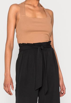 Kobieta ubrana w bezrękawnikowy beżowy prążkowany top oraz wysokowalne czarne spodnie wiązane szerokim materiałowym paskiem w talii.