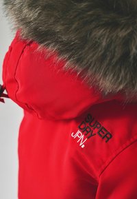 Röd parkas med en huva fodrad med päls. Har svart broderad text "SUPER DRY" och "JPN" på kragen. Slät textur, vattentätt material.