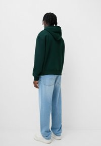 Felpa verde con cappuccio regolabile, abbinata a jeans larghi di un blu chiaro e sneakers bianche, vista da dietro.