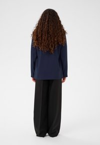 Jūras zila lielgabarīta žakete no gludas auduma, kombinēta ar brīvi pieguļošām melnām biksēm, kurām abām ir minimālistisks dizains un nav redzamu rakstu.