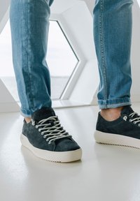Tmavomodré semišové tenisky s off-white gumovými podrážkami a šnúrkami, zobrazené s vyhrnutými svetlomodrými džínsami pred hexagonálnym oknom.