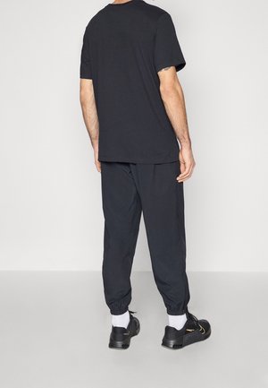 Homme debout, dos à la caméra, portant un t-shirt noir, un pantalon de jogging noir, des chaussettes blanches et des baskets noires.