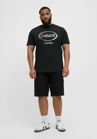 Schwarz über T-Shirt mit einem grafischen Druck in einem kreisförmigen Design, kombiniert mit schwarzen Cargo-Shorts und weißen Sneakern mit schwarzen Akzenten.