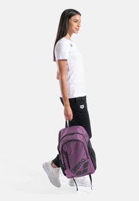 Zaino viola e nero con due cerniere frontali e logo prominente, tenuto da una persona con t-shirt bianca e pantaloni neri, in piedi di lato.