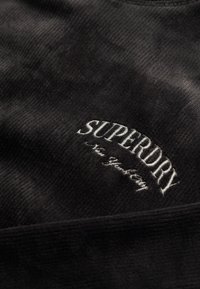 Tejido de pana negra con texto bordado en blanco "SUPERDRY New York City". La textura es rugosa y suave, con un borde liso.