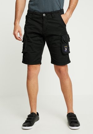 Mand iført sorte cargoshorts med patches og sorte sneakers, stående med den ene hånd i lommen mod en ensfarvet baggrund.