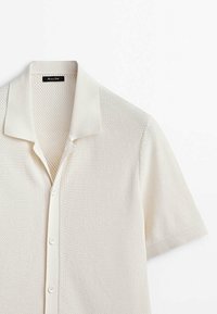 Polo en tricot crème à manches courtes avec motif texturé, fermeture boutonnée devant et col plat, présenté sur fond blanc.