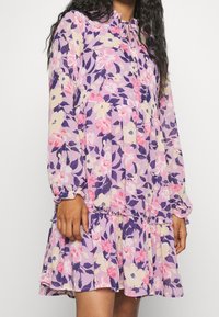 Květinové šaty v odstínech fialové, růžové a krémové. Mají dlouhé rukávy, volánkový lem a zavazování kolem krku. Vyrobeno z lehkého materiálu s hladkou texturou.