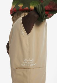 Beige broek met een zakdetail, featuring witte handgeschreven tekst: "Wild heart, Free spirit, Dare to be original" en het jaar "1984."