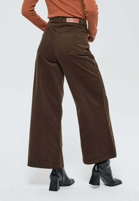 Pantalons larges en velours côtelé marron avec une taille haute, dotés de poches arrière et d'une étiquette rectangulaire sur la taille. Bottines à talons noires.