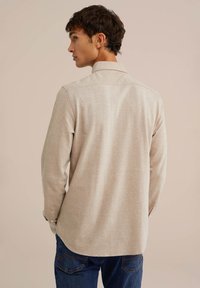 Camisa de manga larga beige con una textura sutil, cuello abotonado y dobladillo redondeado. Combinada con jeans de mezclilla azules, mostrando la vista posterior.