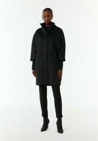 Cappotto di lana nero con collo alto, due tasche frontali e taglio dritto. Indossato sopra pantaloni neri aderenti e stivaletti con tacco.