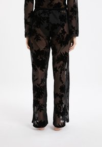 Achteraanzicht van een persoon in zwarte doorschijnende broek en top met bloemenvelvetpatronen, waarbij zwarte onderkleding eronder zichtbaar is.