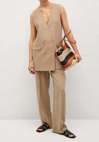 Beige ärmellose Blazer mit doppelreihigen Knöpfen, kombiniert mit lockeren Hosen. Dazu gehört eine gewebte Tragetasche mit Streifen in Braun, Schwarz und Creme.