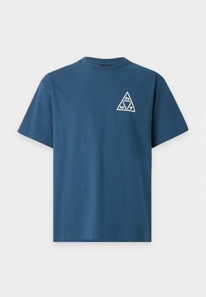 Mężczyzna w niebieskim t-shircie z białym trójkątnym logo zawierającym litery h, u, v, f na plecach, w połączeniu z jasnymi spodniami.