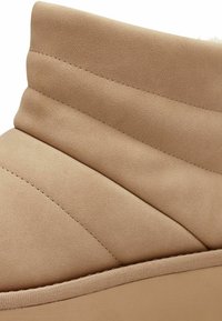Botín beige hasta el tobillo con textura acolchada, parte superior de tela suave, cuello acolchado y detalles de costura a lo largo de los lados, que presenta una suela gruesa.