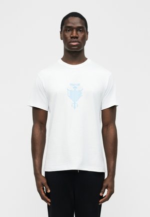 Mies seisoo kasvot eteenpäin, päällään valkoinen Versace Jeans Couture -t-paita, jossa on vaaleansininen logo, ja mustat housut, yksiväristä taustaa vasten.