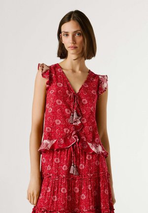 Femme portant une robe rouge à fleurs sans manches avec des volants et des liens à pompons, debout contre un fond clair uni.