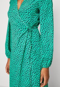 Grünes Wickelkleid mit schwarzem Herzmuster, langen Puffärmeln und einem seitlichen Bindedetail. Hergestellt aus leichtem, geschmeidigem Stoff.