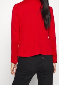 Red fitted blazer with a tailored design, long sleeves, buttoned cuffs, and two side pockets, paired with black pants.